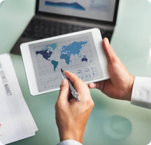Person reviewing a world map and trade documents on a tablet beside a laptop