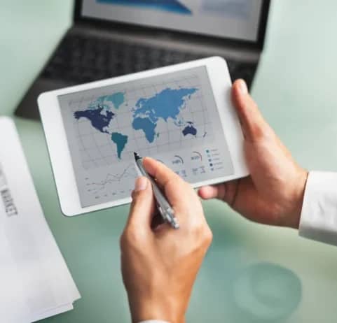 Person reviewing a world map and trade documents on a tablet beside a laptop